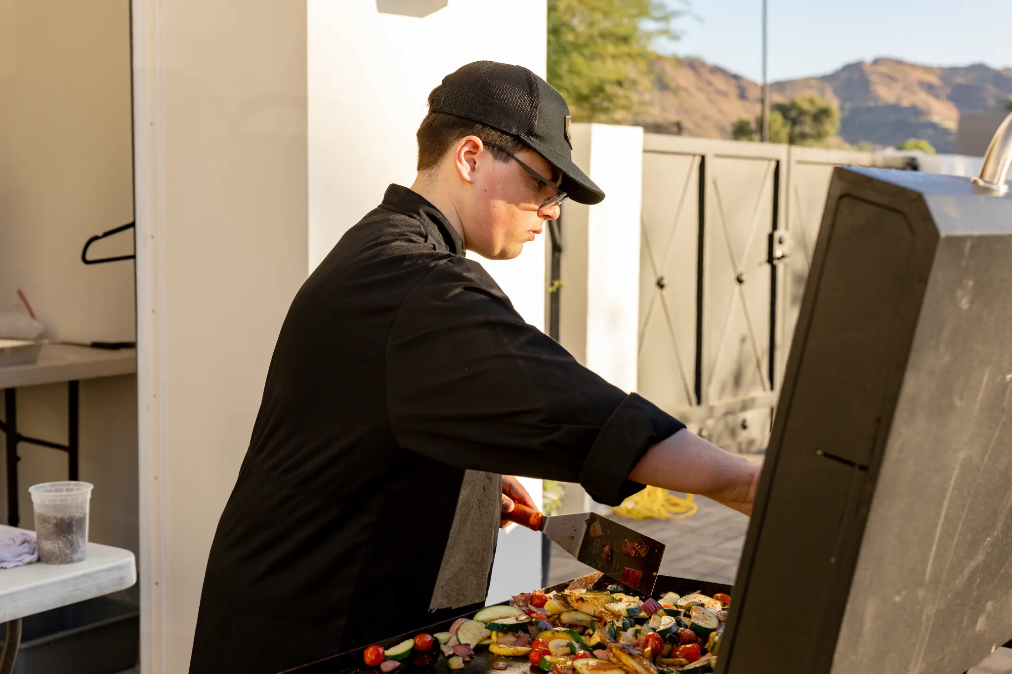 Line cook grilling fresh vegetables on an outdoor grill with Arizona mountains in background