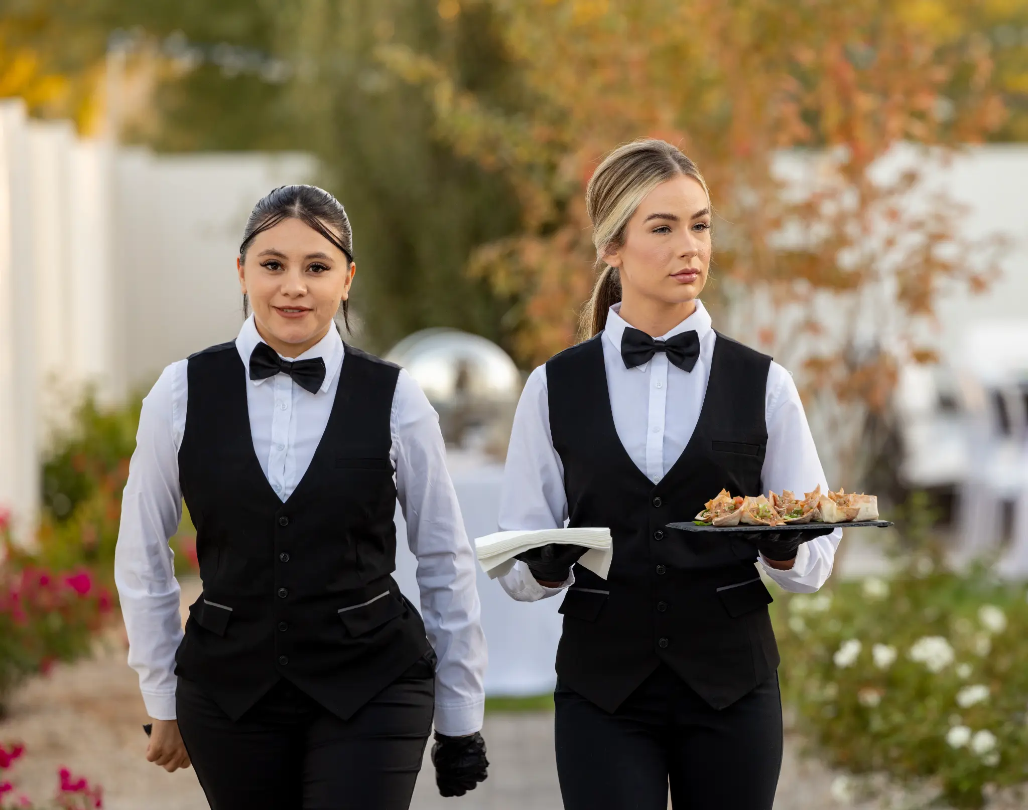 Two professional catering staff in bow ties carrying appetizer trays