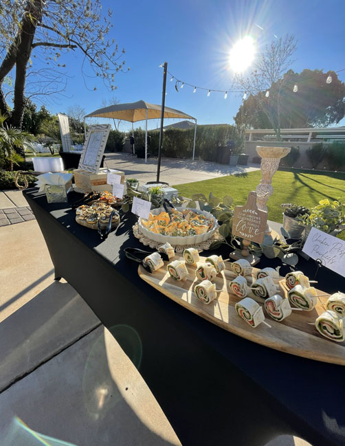 Outdoor appetizer table with bruschetta, roll-ups, and fresh garnishes in sunlight